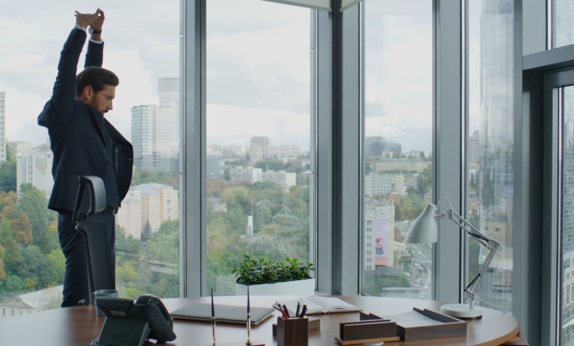 A man in a suit stands with arms raised in front of a large window