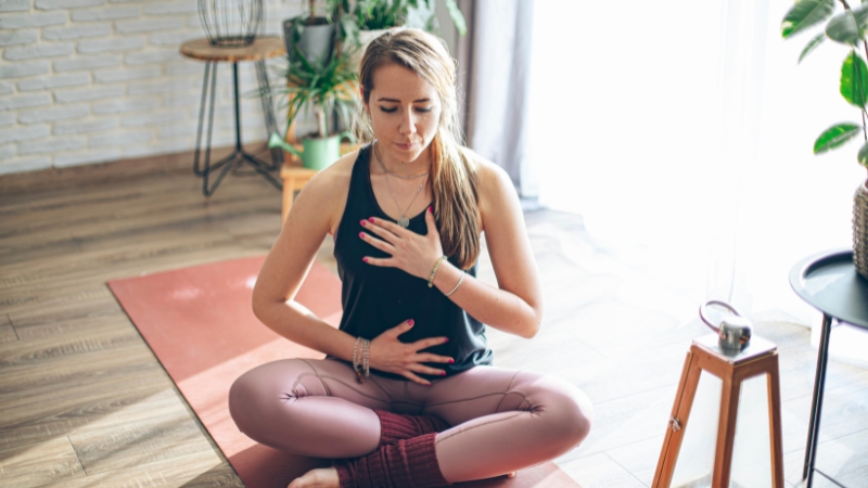 Woman sitting cross-legged on a yoga mat practicing pranayama for anxiety with hands on chest and abdomen