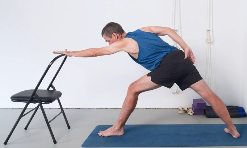 A person in a blue tank top performs a yoga stretch on a mat, extending one arm to a chair for support